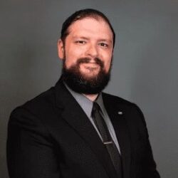 A man with a beard wearing a black suit, dark shirt, and tie poses against a plain, gray background.