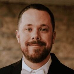 A man with short brown hair and a beard, wearing a tuxedo and bow tie, smiles at the camera against a blurred stone background.