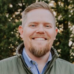 A man with short light hair and a beard smiles outdoors, wearing a green jacket over a blue collared shirt, with trees in the background.