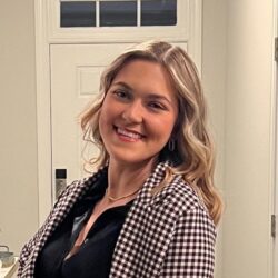 A woman with wavy blonde hair, wearing a black top and a black-and-white checkered jacket, smiles while standing indoors near a white door.