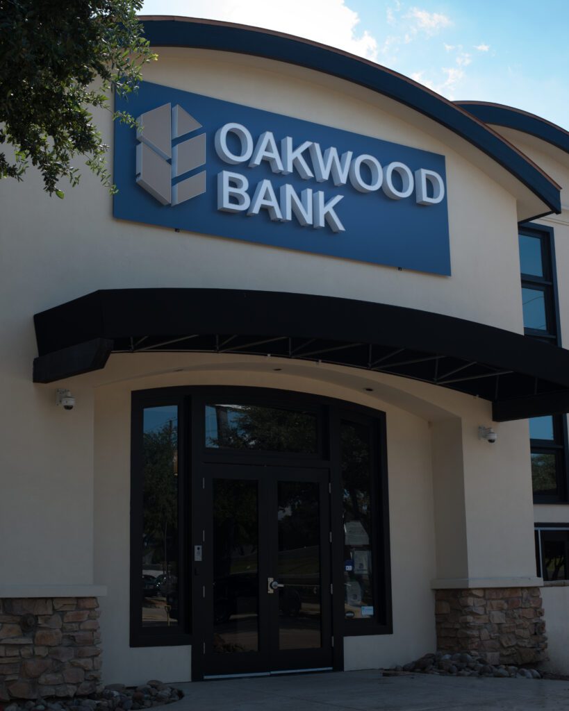 Front entrance of a commercial building with a sign that reads "Oakwood Bank," known for banking M&A, above glass double doors and stone accents on the exterior.