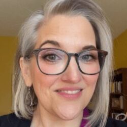 A person with short, grayish-blonde hair and glasses smiles at the camera in a room with yellow walls and shelves in the background.