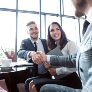 Three professionally dressed people meet in an office, with two of them shaking hands and the third holding a folder, suggesting a business agreement or interview.
