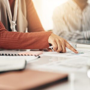 A person wearing a rust-colored sleeve points to a document on a desk covered with papers, notebooks, and folders during a meeting.