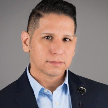 A man with short dark hair, wearing a navy blazer and light blue shirt, poses confidently against a plain gray background, embodying the spirit of digital modernization.
