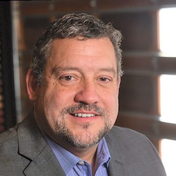A middle-aged man with short curly hair and a goatee, wearing a gray suit jacket and blue shirt, smiles at the camera against a blurred indoor Oakwood Bank background.