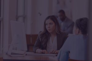 A woman in business attire sits at a desk, typing on a computer and talking to another person; two more people are visible in the background.