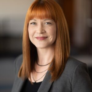 A woman with straight red hair and bangs is wearing a dark blazer and black top, smiling slightly in a blurred indoor setting, possibly at Texas Security Bank.