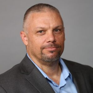 A man with short hair and a goatee wearing a gray suit jacket and light blue shirt poses in front of a plain gray background.