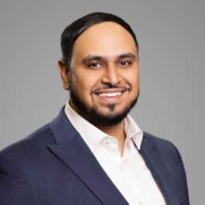 A man with short dark hair and a beard, wearing a navy suit jacket and white dress shirt, smiles at the camera against a plain gray background.
