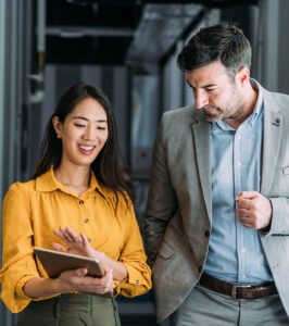 Two professionals walk down a hallway, with the woman in a yellow blouse showing something on a tablet to the man in a grey suit.
