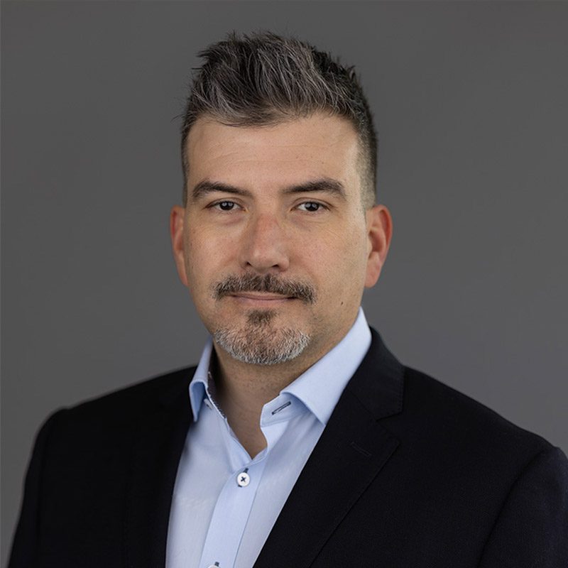 Man with short gray hair and goatee, embodying leadership in a dark suit jacket and light blue shirt, facing forward against a plain gray background.