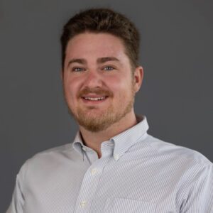 A man with short brown hair and a beard, wearing a light-colored, button-up shirt, smiles at the camera against a plain gray background.