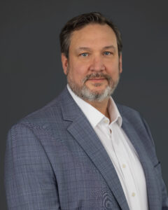 Man with short hair and a beard wearing a light gray plaid suit jacket and white shirt poses in front of a plain dark background.