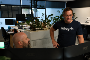 Two men work in a modern office with plants and computer monitors; one stands wearing an “Integris.” shirt, while the other sits at a desk—collaborating on projects powered by CORE Managed AI and IT Services.
