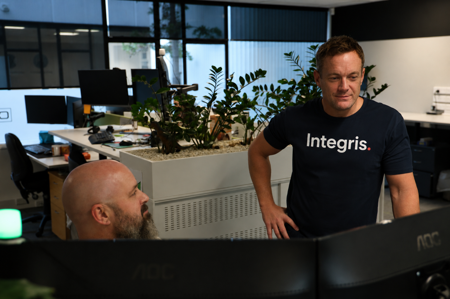 Two men work in a modern office with plants and computer monitors; one stands wearing an “Integris.” shirt, while the other sits at a desk—collaborating on projects powered by CORE Managed AI and IT Services.