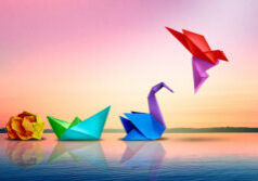 Four colorful origami shapes—a crumpled ball, boat, swan, and flying bird—are arranged on calm water against a pastel sunset sky.