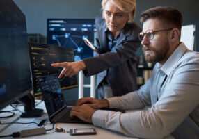 Caucasian businesswoman pointing at cybersecurity dashboard while Caucasian businessman analyzing data on laptop and monitors. Ideal for IT teamwork, network security, risk management