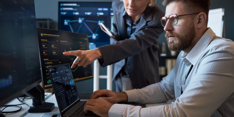 Caucasian businesswoman pointing at cybersecurity dashboard while Caucasian businessman analyzing data on laptop and monitors. Ideal for IT teamwork, network security, risk management