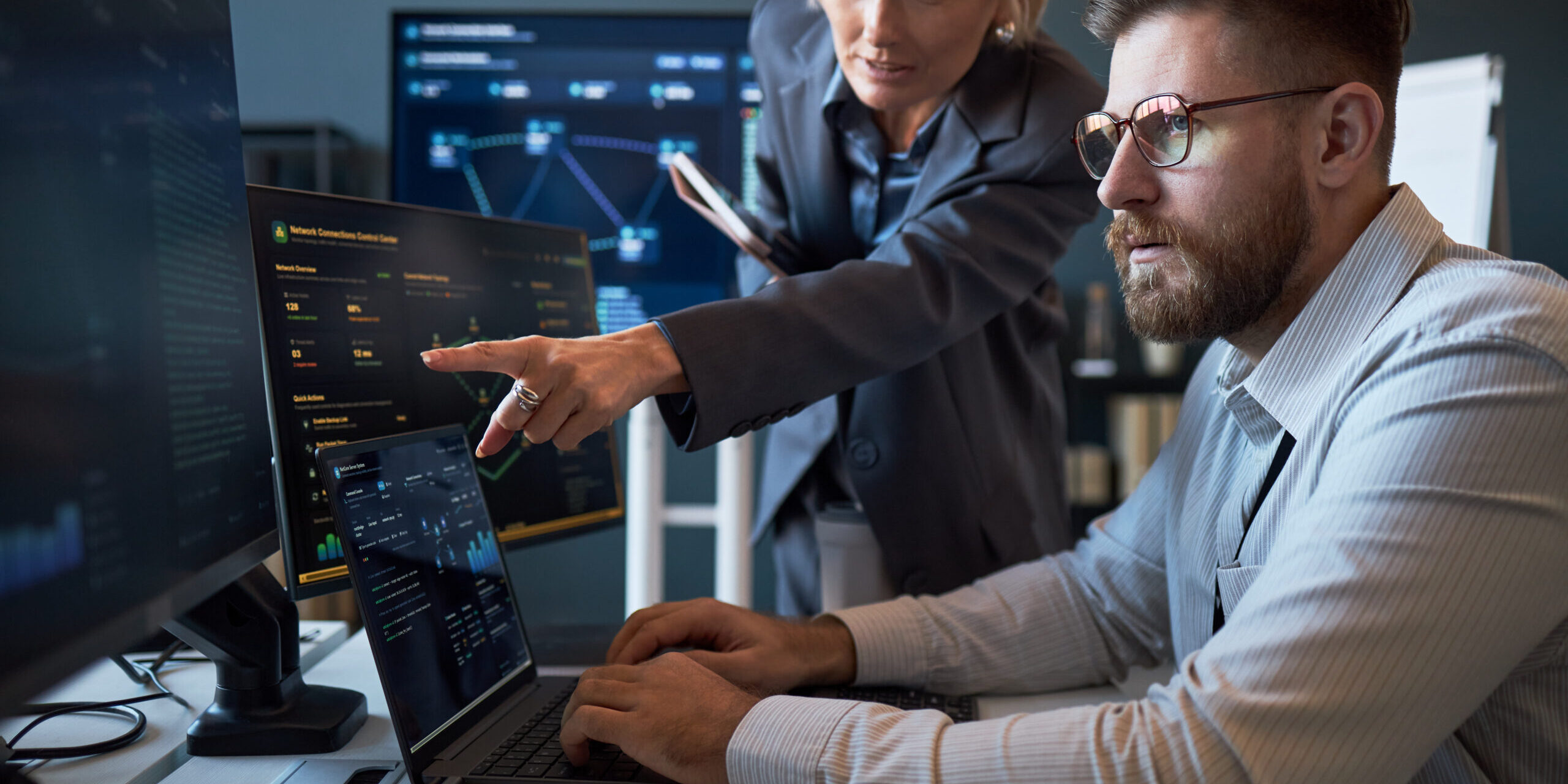 Caucasian businesswoman pointing at cybersecurity dashboard while Caucasian businessman analyzing data on laptop and monitors. Ideal for IT teamwork, network security, risk management