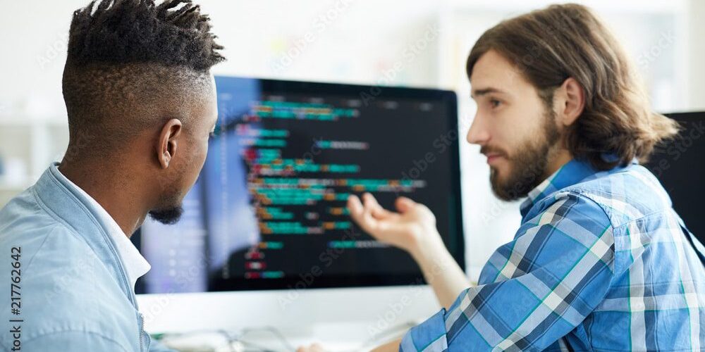 Two men engaging in a discussion while looking at computer code on a monitor.