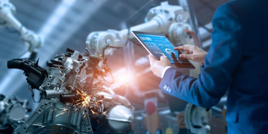 A person in a blue suit operates a tablet in an industrial setting, monitoring robotic arms working on machinery with sparks visible. In this high-tech environment, manufacturers need managed IT services to ensure seamless operations.