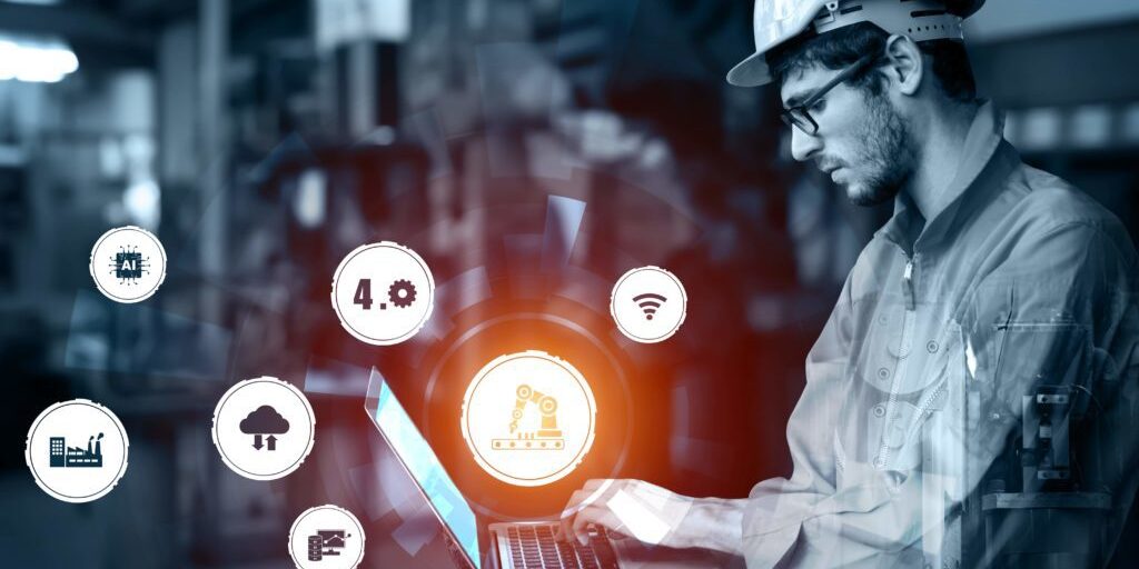 A person wearing a hard hat and safety glasses works on a laptop in an industrial setting. Various technology icons, such as automation and wireless, surround the image.