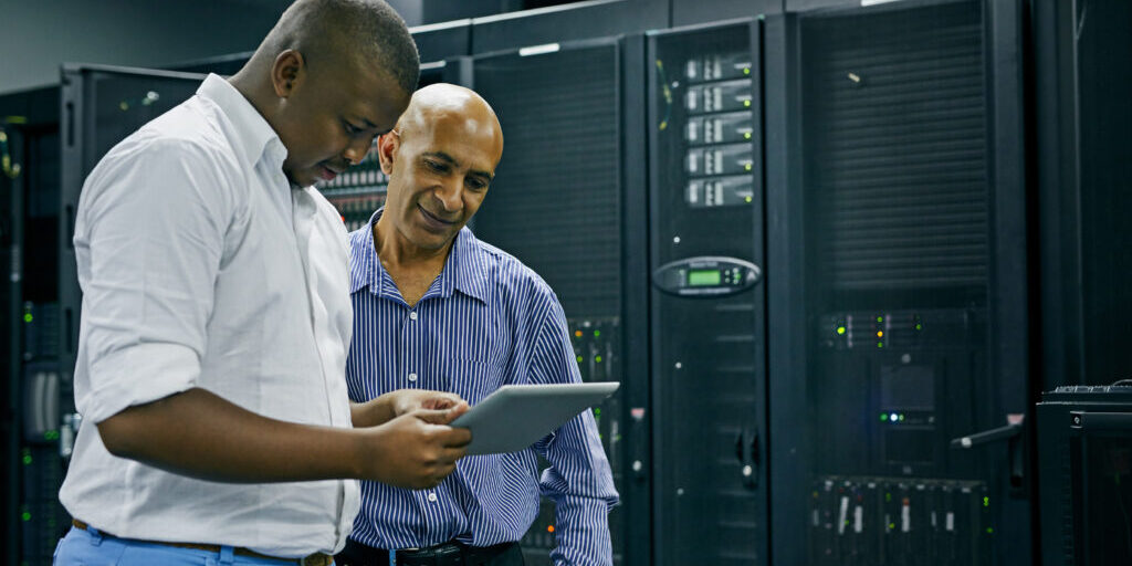 Two men standing in a server room, one holding a digital tablet while the other looks on, surrounded by server racks and computer equipment.