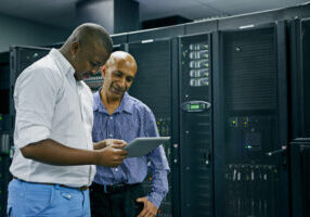 Two men standing in a server room, one holding a digital tablet while the other looks on, surrounded by server racks and computer equipment.