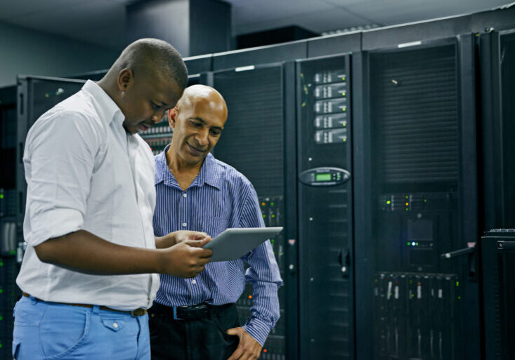 Two men standing in a server room, one holding a digital tablet while the other looks on, surrounded by server racks and computer equipment.