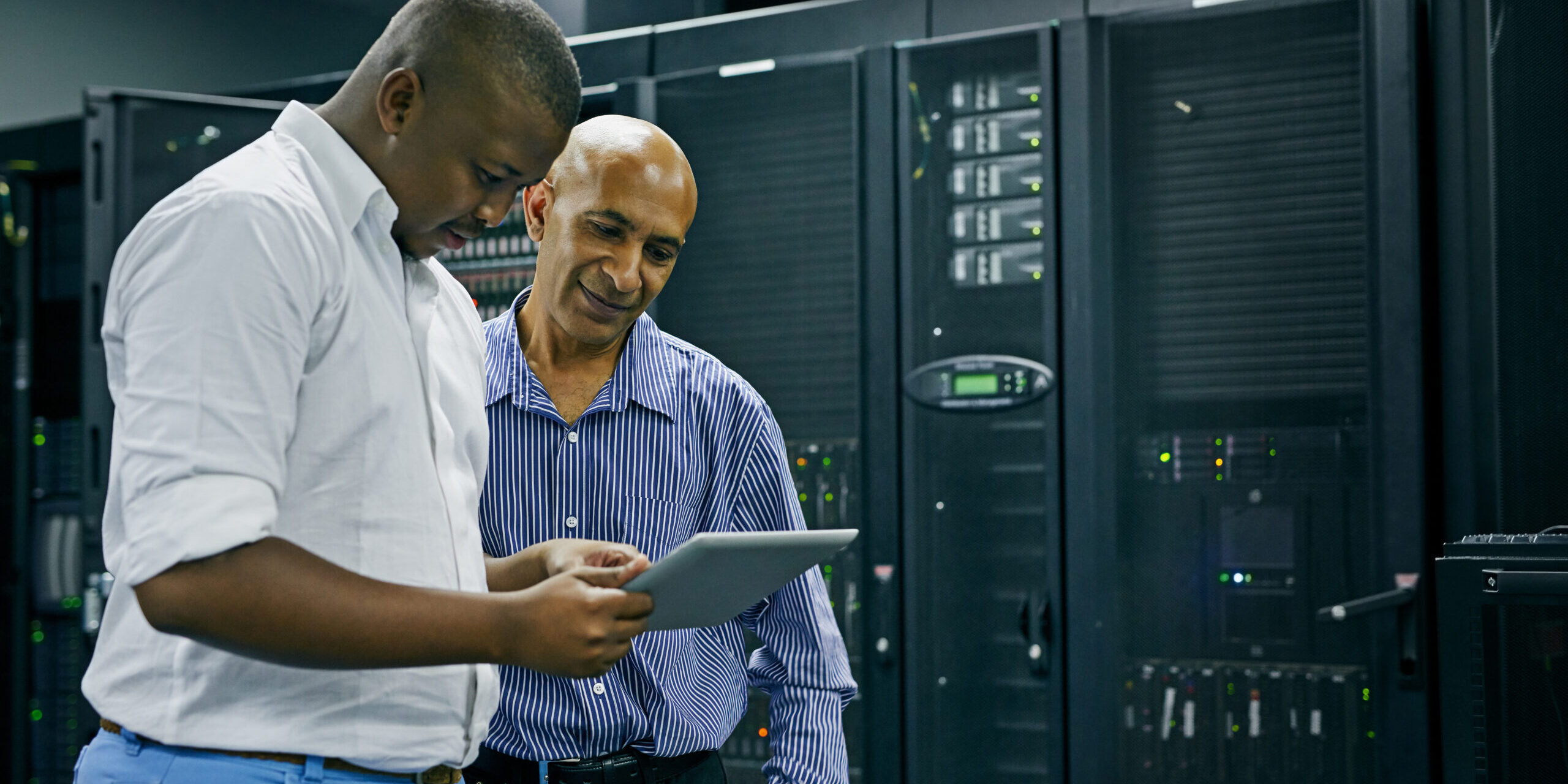Two men standing in a server room, one holding a digital tablet while the other looks on, surrounded by server racks and computer equipment.