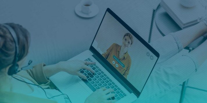 A person wearing a headset is sitting with their feet up on a table, using a laptop to have a video call with another person displayed on the screen. A cup of coffee sits on a nearby table.