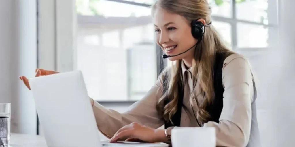 A woman wearing a headset sits at a desk, smiling at her laptop screen, in a bright office with large windows.
