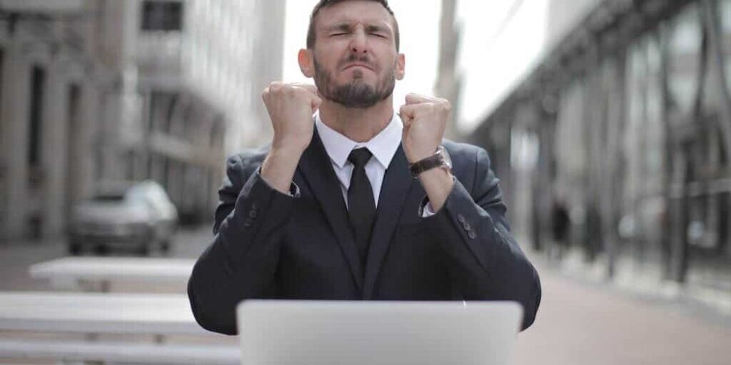 A man in a suit sits outdoors at a table with a laptop, clenching his fists and appearing to celebrate success.