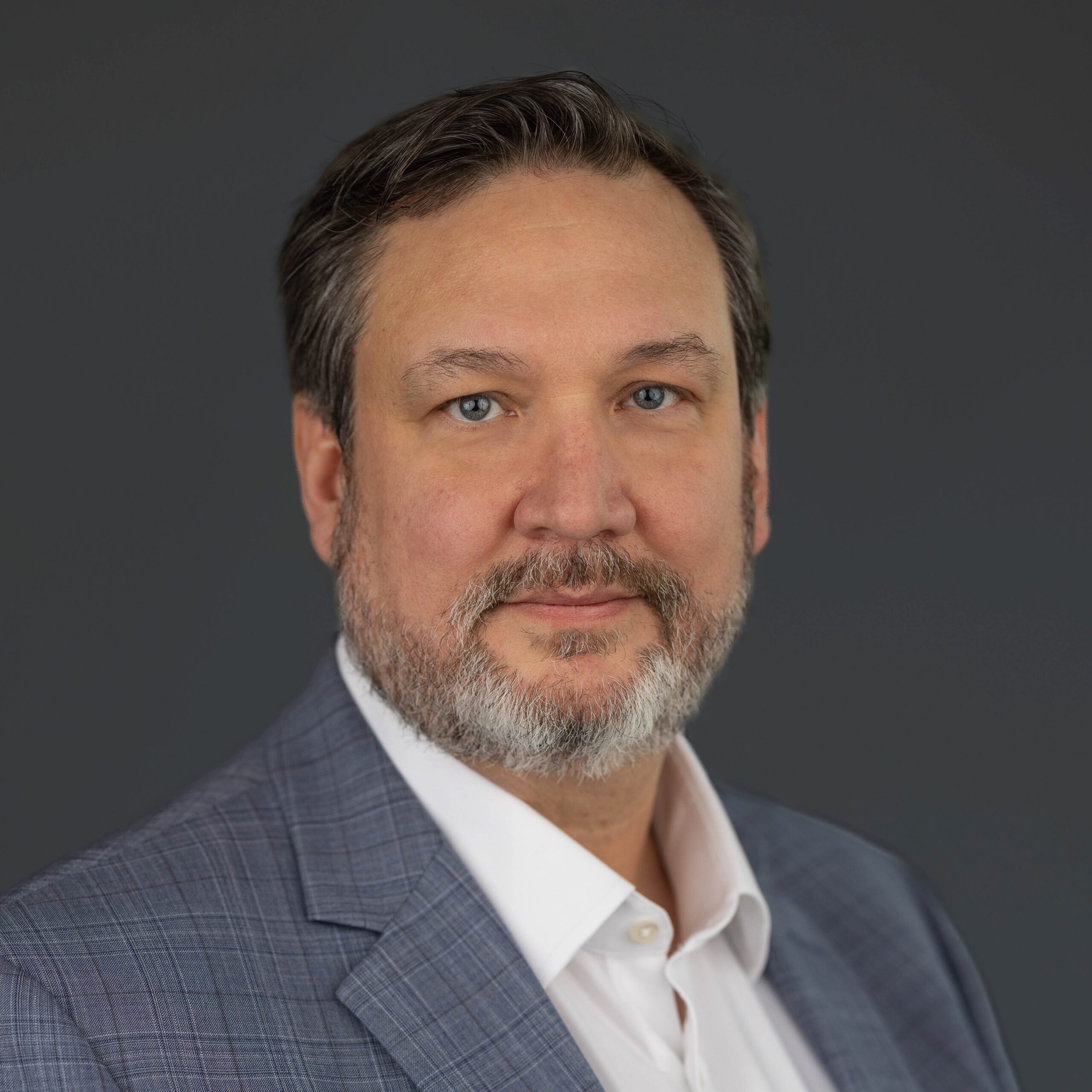 A man with short dark hair, a beard, and a mustache, wearing a white shirt and plaid gray blazer, poses against a plain dark background.