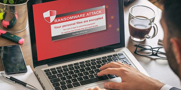 A laptop screen displays a ransomware attack message indicating that personal files are encrypted and a ransom must be paid to recover them.