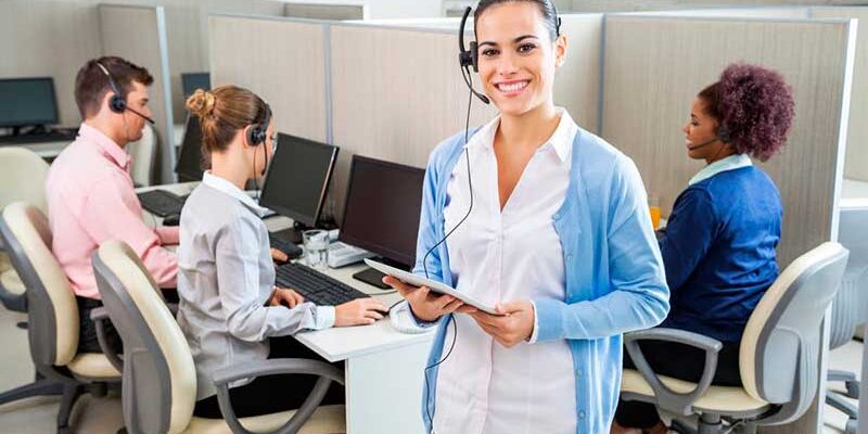A smiling woman wearing a headset and holding a tablet stands in the foreground of an office with other employees working at computer stations.