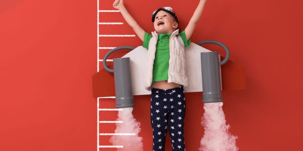 A child wearing a toy rocket backpack and star-patterned pants raises their arms joyfully against a red wall with a chalk-drawn height chart.