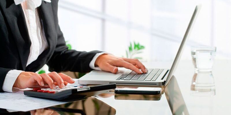 A person in formal attire works on a laptop and uses a calculator at a glossy desk with a glass of water and documents nearby.
