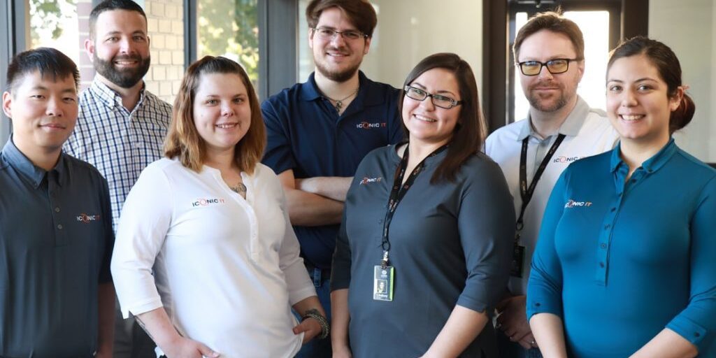 A group of seven people posing for a photo indoors, wearing casual business attire and nametags, standing in front of large windows and a door.