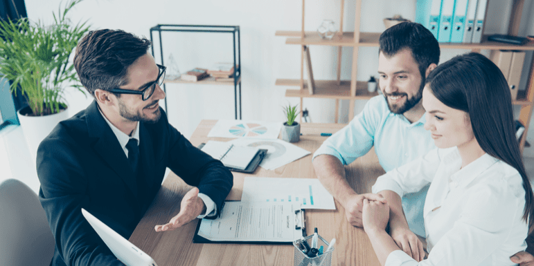 Three people are sitting at a desk with documents and a tablet. A man in a suit is talking to a couple who are sitting across from him, smiling. Shelves with files and plants are in the background.