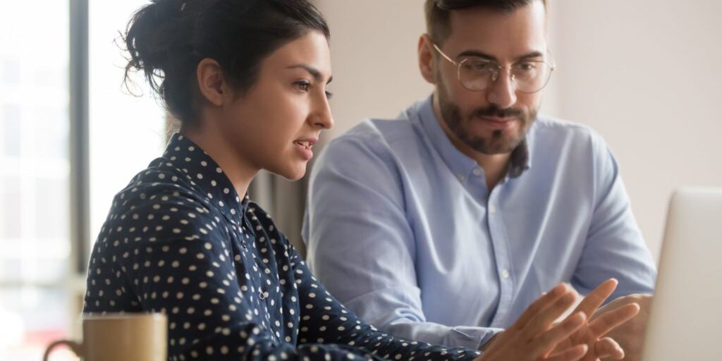 Two colleagues sit together at a desk, looking at a laptop screen. The woman points at the screen while the man observes. A coffee mug is placed on the desk beside them.