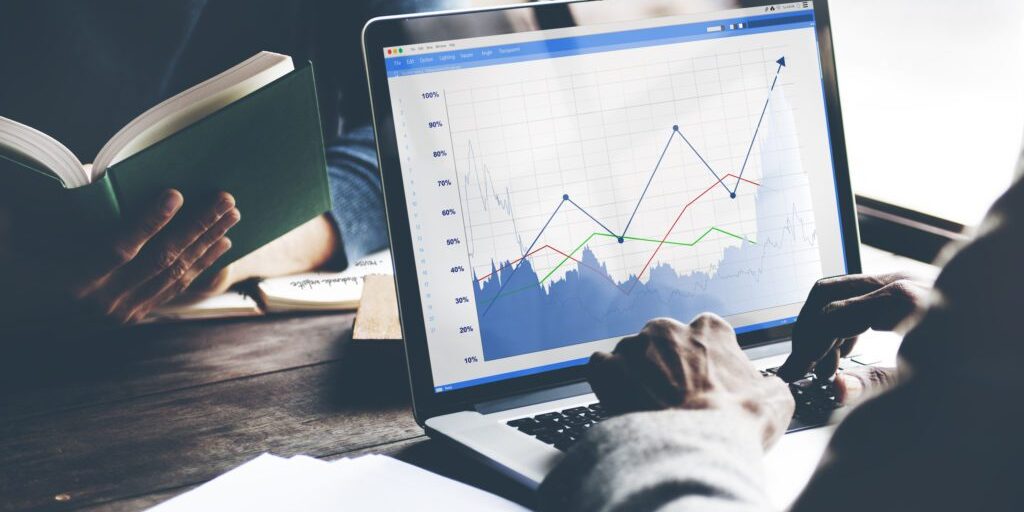 Two people are working together. One is reading a book and the other is using a laptop displaying a graph with multiple colored lines and data points.