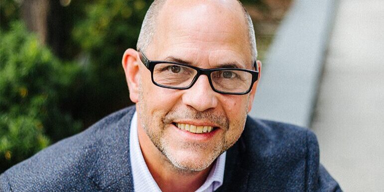 A man with glasses and a beard smiles while wearing a dark blazer and light-colored shirt outdoors.