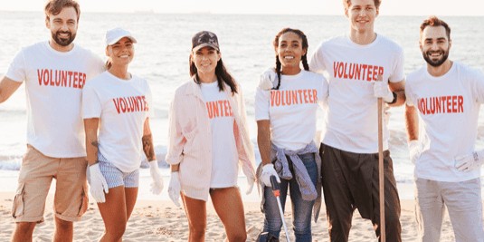 A group of six volunteers in matching shirts and gloves stands together on a beach, smiling. Some hold trash bags and a litter pick, suggesting they are participating in a beach clean-up event.