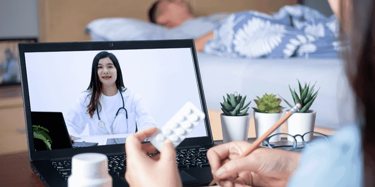 A person holds a blister pack and writes notes while having a telemedicine consultation with a doctor on a laptop. A bed and various plants are in the background.