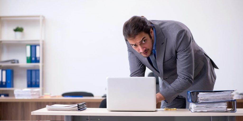 A man in a gray suit leans over a desk, looking at a laptop. On the desk are tall stacks of papers. The background features shelves with folders and office supplies.