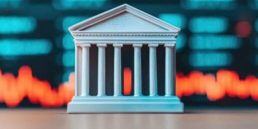 A model of a classical bank building with columns is in front of a blurred background showing red and blue financial stock market data.