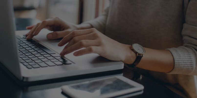 A person types on a laptop keyboard with a smartphone placed nearby on a dark surface.