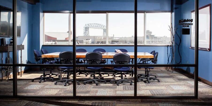 A glass-walled conference room with a wooden table surrounded by ten chairs, offering a window view of the cityscape. A sign on the right reads "Bulldog Conference Room," perfect for meetings or discussions about IT support in Duluth.